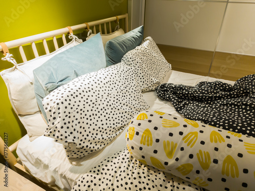 Cozy bedroom interior with colorful pillows and patterned bedding on an unmade bed. Modern home design with playful polka dots and bright yellow accents creating a cheerful, relaxed atmosphere.
