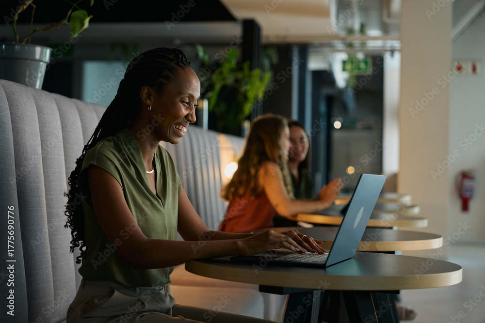 Fototapeta premium Side view of smiling young black businesswoman in smart casual using laptop at desk in office lobby