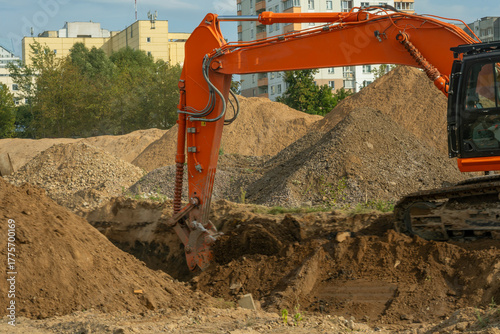 Excavator digging soil on construction site with urban buildings in the background. Industrial excavator performing earthmoving operations at construction site for residential infrastructure.