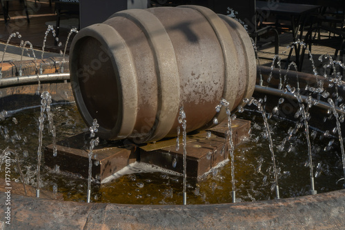 Water fountain with wooden barrel and circular jets in outdoor setting. Rustic barrel fountain with metal stand and water jets in public plaza with tables and chairs under natural daylight.