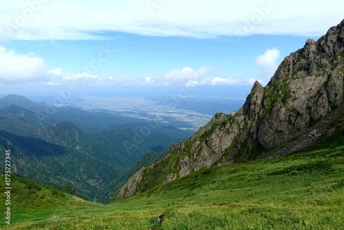 Jagged rock spires frame a sweeping view from Mount Ashibetsu, Hokkaido, across green alpine slopes to distant plains under a bright summer sky.