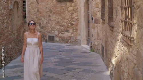 Elegant woman exploring an old italian town. Young tourist woman in a summer dress walking through a narrow cobblestone street in an ancient italian town, admiring the medieval architecture in tuscany