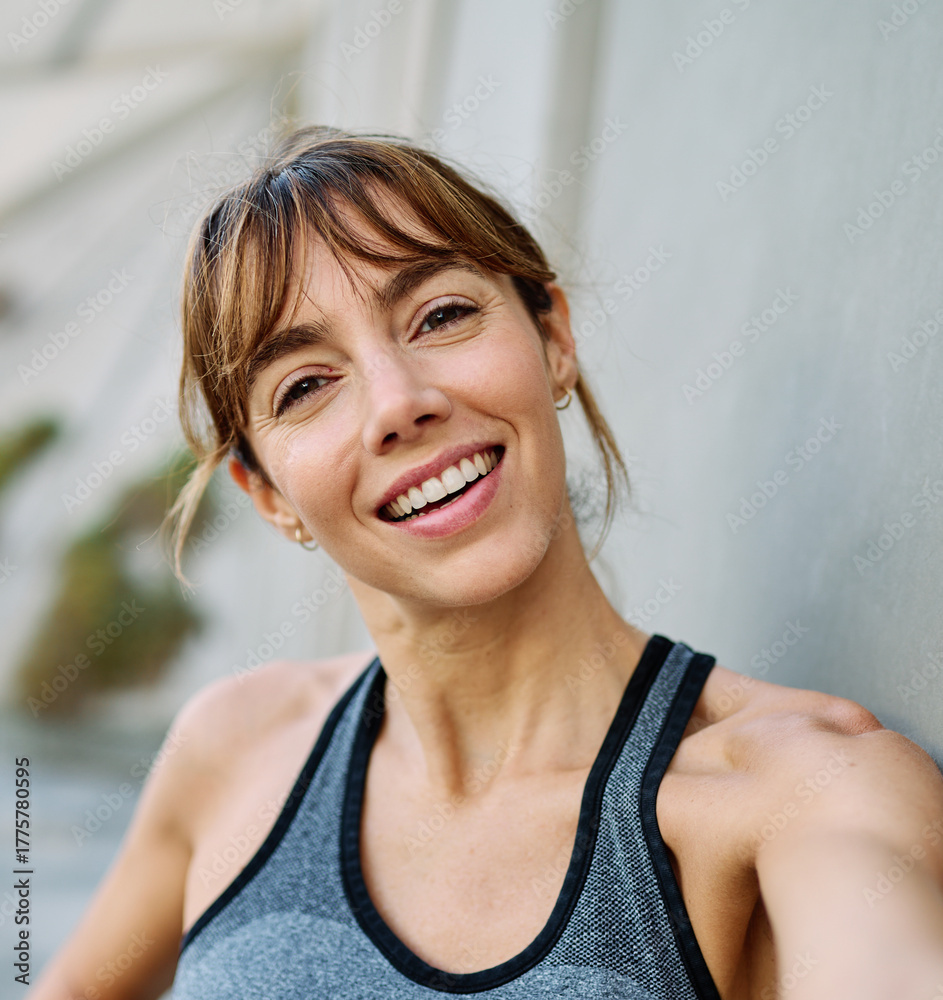 Fototapeta premium Young woman taking a selfie while posing against a wall after workout in athletic wear at urban location
