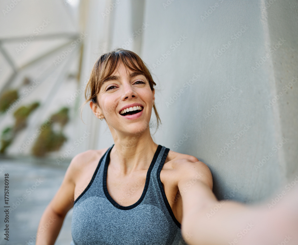 Fototapeta premium Young woman taking a selfie while posing against a wall after workout in athletic wear at urban location