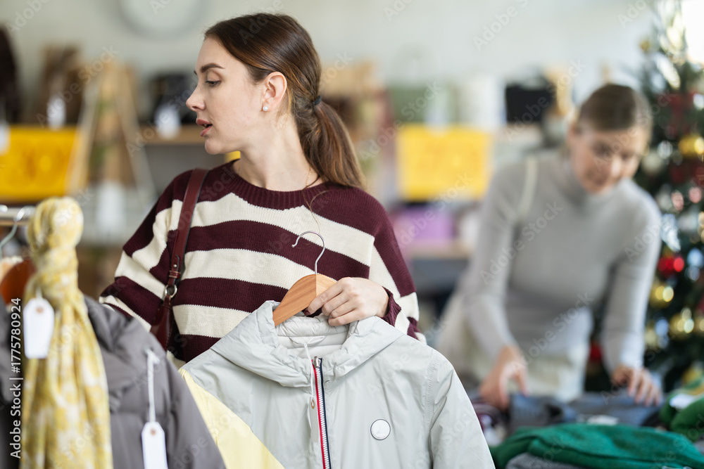Fototapeta premium Adult woman stands in a store decorated for the new year and chooses outerwear among jackets and coats.