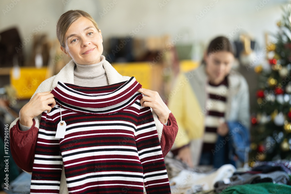 Fototapeta premium Positive young woman choosing some sweater in clothing shop during Christmas sale