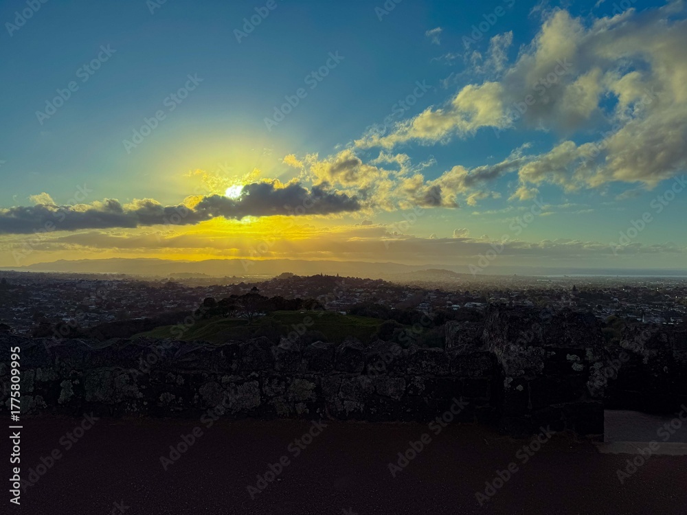 Obraz premium The sun is setting into the horizon at the city. The view is from high ground green mountain in summer time. Beautiful orange sunset view of Auckland city.