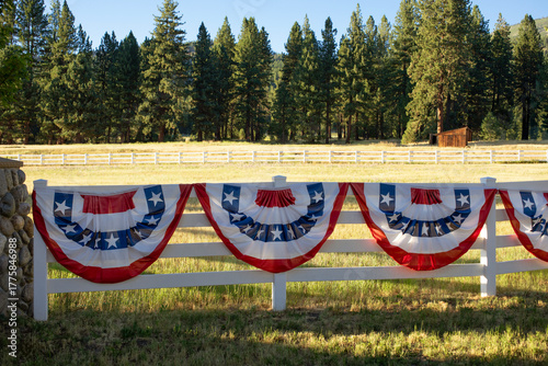 Red, white, and blue patriotic bunting with white stars on the blue section, shaped in semicircles and draped along a fence for Fourth of July decoration. A slice of Americana in a rural area.
