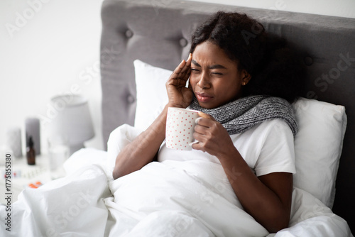 Foto Sad young black lady lies in bed wrapped in a scarf, holding a cup of hot drink