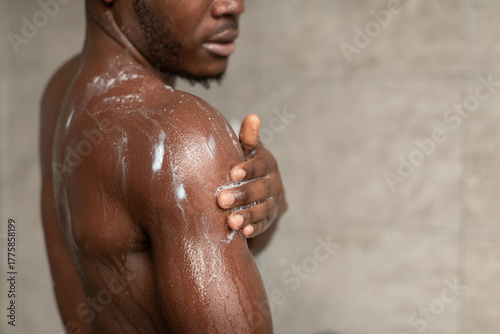 Unrecognizable Black man stands in an indoor bathroom while washing his body with shower gel. He is focused on his hygiene routine, enjoying the relaxing moment.