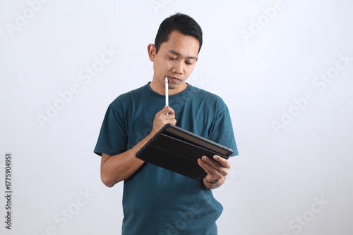 Thoughtful Asian man working on digital tablet with stylus pen on white background.