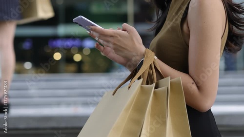 Female tourist walking city streets, carrying multiple shopping bags while browsing smartphone, experiencing digital retail therapy with evident enjoyment and urban lifestyle satisfaction.