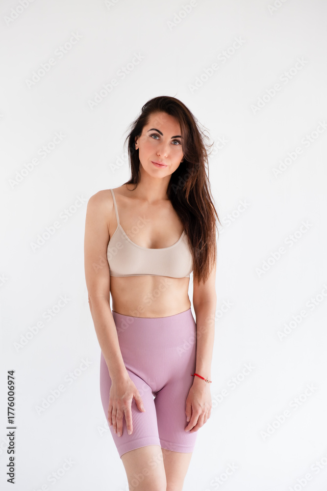 Fototapeta premium Sports woman standing in activewear with abs muscles, looking at camera isolated on white background. Caucasian 30s fitness girl wears beige top, pink leggings. Happy fit smiling brunette, blue eyes.