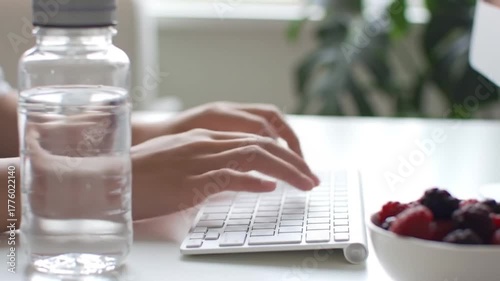 A person typing on a computer keyboard with a healthy snack of fresh berries and a bottle of water on the desk.