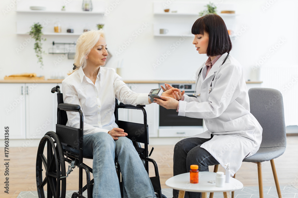Fototapeta premium A female doctor measures the blood pressure of an elderly woman in a wheelchair during a home visit, with medicine on a table.