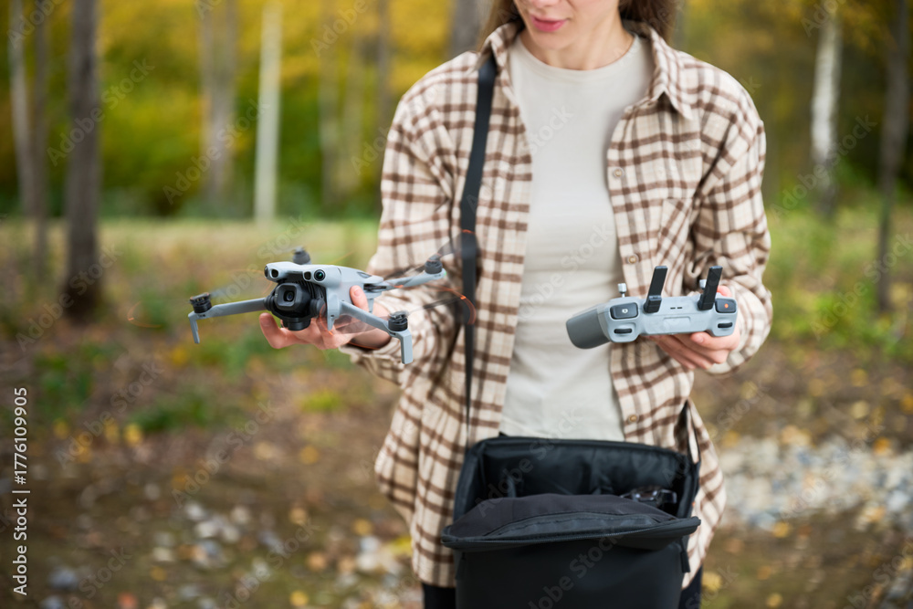 Fototapeta premium A woman stands in a vibrant forest, holding a ready-to-fly drone in one hand and its controller in the other. She enjoys the colorful autumn scenery.