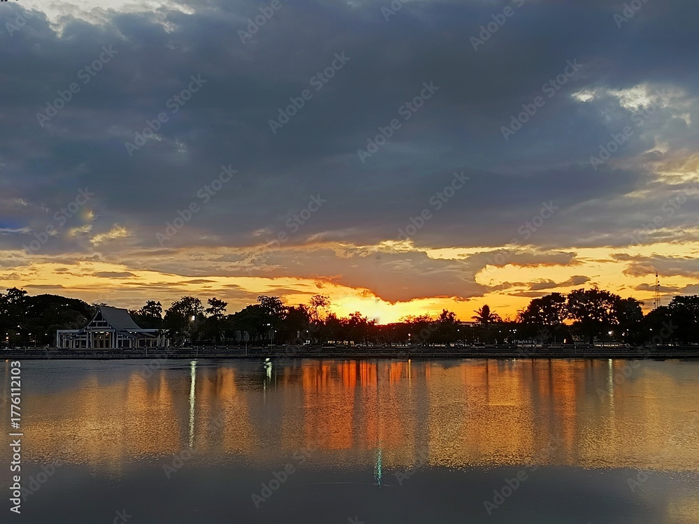 Obraz premium Golden Sunset over Lake: A tranquil lake mirrors a breathtaking sunset, its waters reflecting the fiery hues and silhouetted trees along the horizon, creating a scene of serenity.