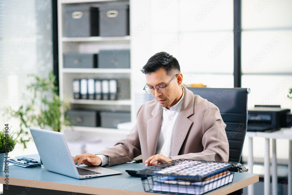 Obraz premium Confident Asian businessman standing at desk, reviewing report and working on laptop in modern office