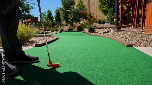Playing mini golf with a yellow ball and orange putter on green astroturn on a sunny day - shot from behind golfer