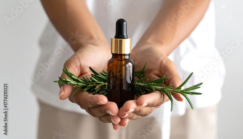A person holds a brown glass bottle with gold cap, rosemary sprigs in hands
