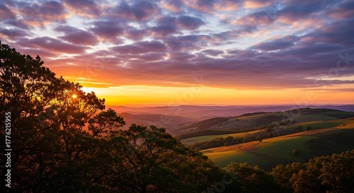 Golden hour sunset paints a tranquil rolling landscape. Sky bursts with vibrant oranges, purples, yellows. Picturesque evening vista over hills, silhouetted trees
