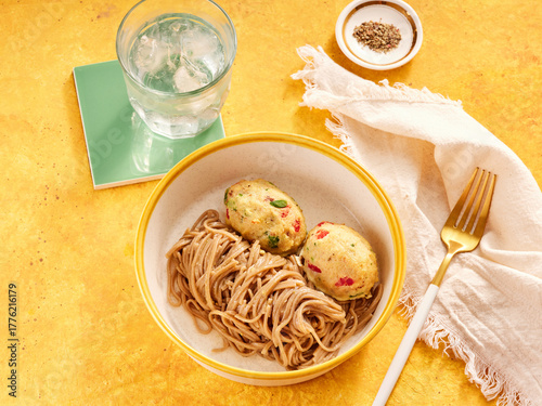 A bowl filled with noodles sits beside two colorful dumplings on a cheerful yellow table. A glass of cold water and a napkin complete the setting, inviting a satisfying meal