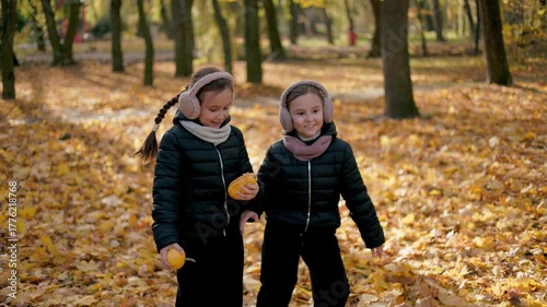 Two twin sisters dance and play while listening to music in the park.The autumn sun breaks through the sparse branches of the trees, illuminating a lawn covered with a carpet of yellow, red and orange