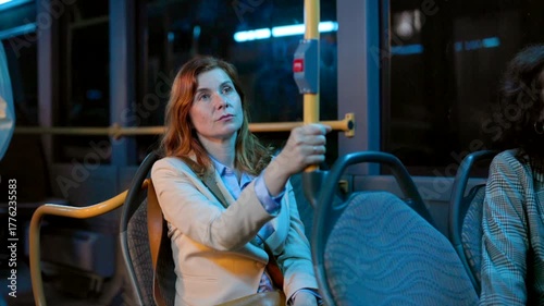 Pensive and tired businesswoman traveling home by public transport at night, feeling sleepy and exhausted after a long day at work, holding onto a handrail while commuting on the bus