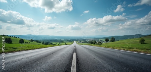 Fototapeta Naklejka Na Ścianę i Meble -  Empty asphalt road curves towards distant valley and small town under cloudy sky. Green rolling hills and scattered trees frame the expansive landscape and horizon.