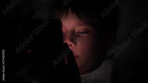 In the dark, a Caucasian boy of 11-12 years old uses a gadget in bed while lying in bed. A child with a phone in bed at night, his face illuminated by the light of the gadget.