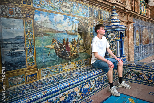 Young man sitting beside the colourful ceramic tiles representing the Canary Islands at Plaza de España in Seville