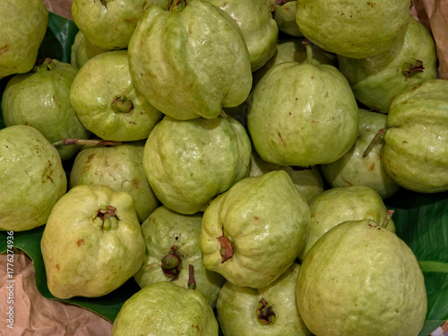 A close-up view of a large, dense pile of whole, light green guavas, resting on green leaves and brown paper.
