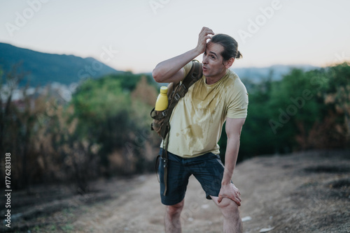 A man with a backpack and water bottle pauses on a dirt trail, catching his breath after a strenuous climb. Warm light and greenery frame a moment of stamina and perseverance.
