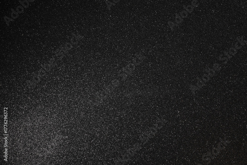 Black glitter texture with fine granular relief, full surface in sharp focus while subtle scattered sparkles and soft light glow form abstract depth