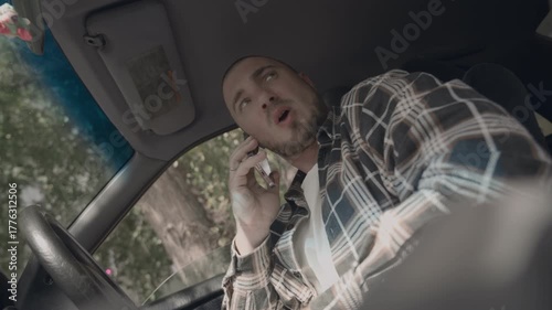 A man is sitting in his car, talking on the phone while driving. He is a hipster businessman making a business call from his vehicle.
