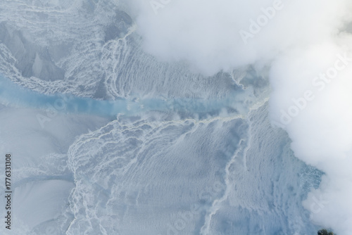 Aerial view of a surreal, milky-blue river cuts through a stark, white geothermal landscape, vaporous clouds swirling above, Geothermal Areas, Iceland.