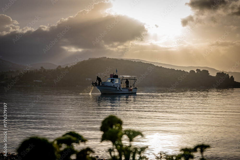 Fototapeta premium The Autumn sun breaking through the clouds casting sun rays on the still sea with a small fishing boat and fisherman checking their catch.