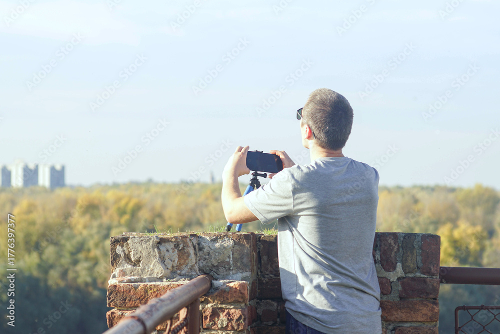 Fototapeta premium Man mounting smartphone on octopus tripod at scenic overlook with city horizon. Represents timelapse recording setup, hands-free filming solution, travel vlogging equipment, and mobile journalism tool