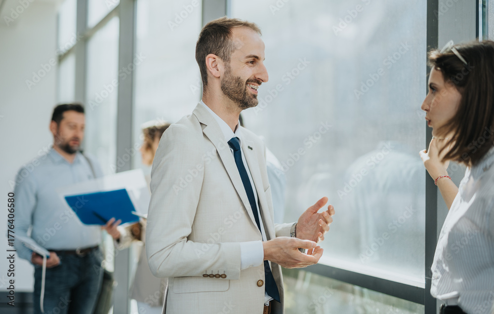 Fototapeta premium A smiling professional in a light suit chats with a colleague near a large window while others review documents in the background, capturing workplace collaboration and networking.