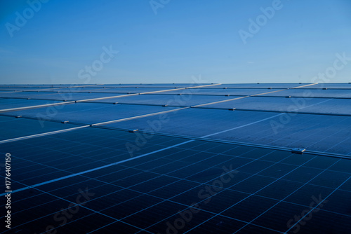 A large solar panel array is seen from above. The panels are blue and are arranged in a grid pattern. The sky is clear and the sun is shining brightly. Concept of clean energy and sustainability