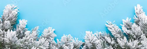 Green artificial fir branches covered with hoarfrost and white snow on light blue table background. Pastel color. Closeup. Wide banner. Empty place for text. Top down view.