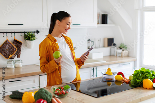 Smiling pregnant woman standing in modern kitchen holding apple in one hand and using cellphone with the other, checking social media feed while cooking or eating
