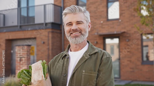 Mature Caucasian man with gray hair and beard holding grocery bag turning head and smiling. Looking directly at camera while standing outside modern residential brick house. Glancing at camera.
