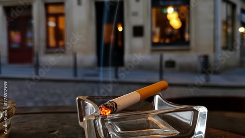 A smoldering cigarette in a glass ashtray rests on a table outside a city building, smoke curling upwards. Nightlife vibes and city glow.