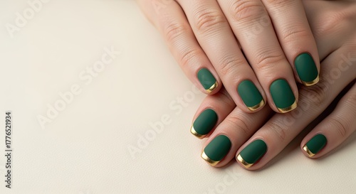 Close-up of manicured hands with green matte nails and gold tips. The background is a soft, neutral color, emphasizing the nail art design.