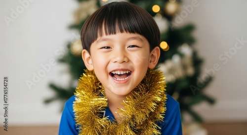 Close-up of a smiling Asian boy wearing a gold tinsel garland, with a softly blurred Christmas tree and warm bokeh lights in the background, bright festive portrait