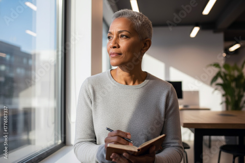 A head-and-shoulders portrait of a mature Black woman with short, gray hair, standing by a large, sunny office window.