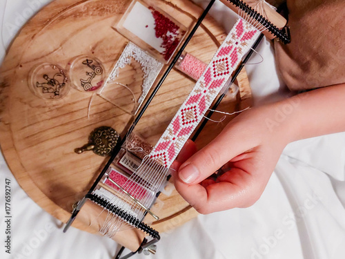 Weaving a bracelet on a loom with a pattern of national Ukrainian motifs in pink tones, creating jewelry from beads as a hobby