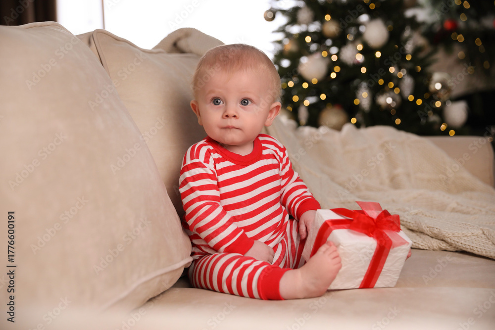 Naklejka premium Cute baby in bright Christmas pajamas holding gift box on sofa at home