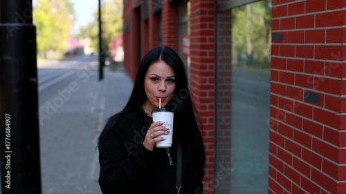 Outside portrait of beautiful attractive brunette young woman drinking juice through straw looking at camera. Stylish girl walking across the city. Positive lifestyle. Happy day. Healthy nutrition. 